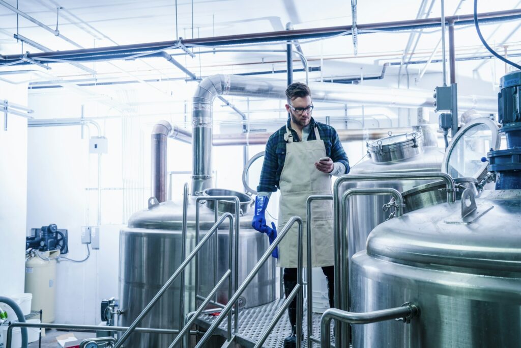 Young man in brewery wearing apron and protective gloves looking at smartphone
